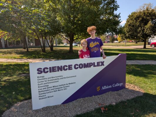  Two young boys are displayed behind a sign reading “SCIENCE COMPLEX.” The boy on the left is wearing a red shirt, while the boy on the right is wearing a dark purple shirt with “Briton” on it in gold lettering. 