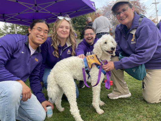 Four adults wearing purple sweatshirts crouch on grass around a white poodle mix dog, all smiling at the camera. Behind them are a purple tent and a few people standing.