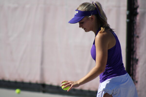 A female tennis player in a purple outfit and visor prepares to serve on a sunny court, holding a tennis ball with focus and determination.