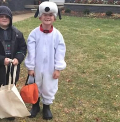 Two boys stand in front of a house holding bags. From left to right, they are dressed as a sci-fi villain in a black cape and armor, and the other as a cartoon dog with black floppy ears.