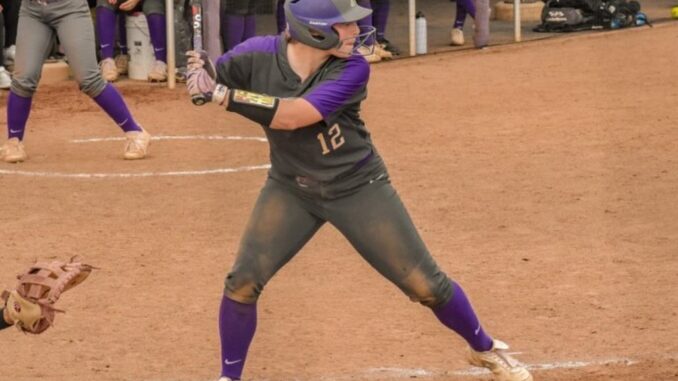 A woman wearing a purple and gray softball uniform stands with a baseball bat held over her shoulder, ready to swing. She is standing in the batter’s box on a softball field.