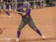 A woman wearing a purple and gray softball uniform stands with a baseball bat held over her shoulder, ready to swing. She is standing in the batter’s box on a softball field.