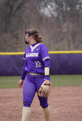 A woman wearing a purple softball uniform walks across a softball field as she looks over her shoulder. 