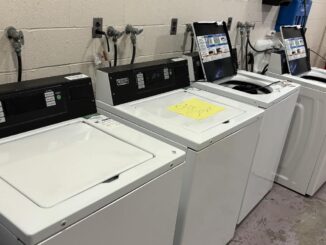 A row of top-loading washing machines, some open and others closed. One of them has a sign on it that reads ‘out of order.’