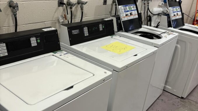 FEATURE A row of top-loading washing machines, some open and others closed. One of them has a sign on it that reads ‘out of order.’