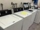 A row of top-loading washing machines, some open and others closed. One of them has a sign on it that reads ‘out of order.’