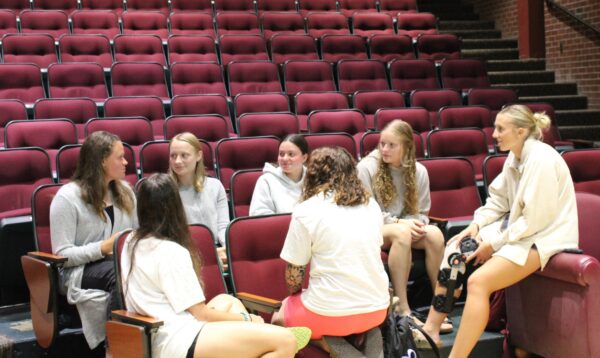 A group of female students scattered across two rows of auditorium style seating are arranged in a circle. The group is engaging in small group discussions.