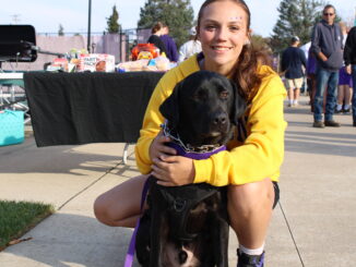 A young woman in a yellow sweatshirt crouches on a sidewalk, her arms around the black lab sitting in front of her. Behind them are tables, a fence and people walking around.