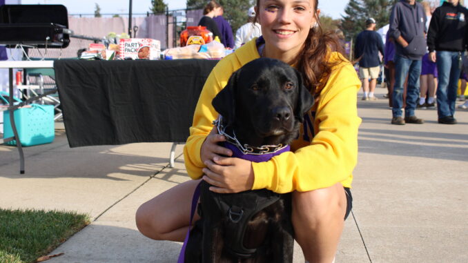 A young woman in a yellow sweatshirt crouches on a sidewalk, her arms around the black lab sitting in front of her. Behind them are tables, a fence and people walking around.