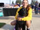A young woman in a yellow sweatshirt crouches on a sidewalk, her arms around the black lab sitting in front of her. Behind them are tables, a fence and people walking around.