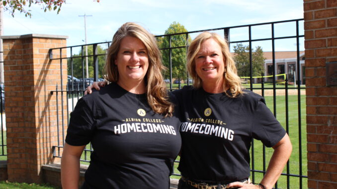 FEATURE Two middle-aged women stand with their arms around each other, smiling at the camera as the sun shines on them. They both wear black T-shirts that read “Albion College Homecoming.” They are outdoors, standing in front of a black fence with brick pillars; in the background a building, trees and cars are visible.