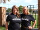 Two middle-aged women stand with their arms around each other, smiling at the camera as the sun shines on them. They both wear black T-shirts that read “Albion College Homecoming.” They are outdoors, standing in front of a black fence with brick pillars; in the background a building, trees and cars are visible.