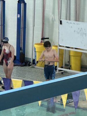 A young boy is displayed. He is standing alongside a community pool edge and adjusting a set of swim goggles. The boy is shirtless and wearing navy blue swim trunks. In the foreground is a set of triangular banners, purple and gold. 