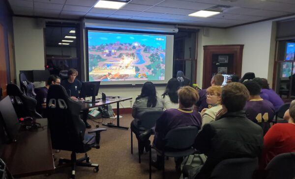 Two people sit in front of a computer at a long table at the frog of a large room with white walls and several windows. Beside them is a projector screen with a video game playing, and several rows of young people in chairs face the image.