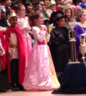 A young girl with her side to the camera stands on a stage. The girl is surrounded by other costumed kids and wearing a pink princess dress. 