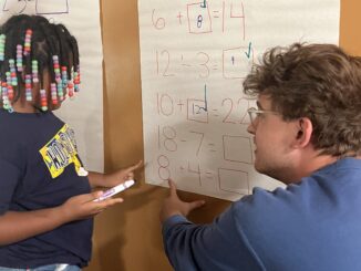 A young man with short brown hair kneels and faces a poster paper. To the left of him is a young girl wearing box braids with beads, also facing the poster paper. On the paper itself is a series of addition and subtraction problems.