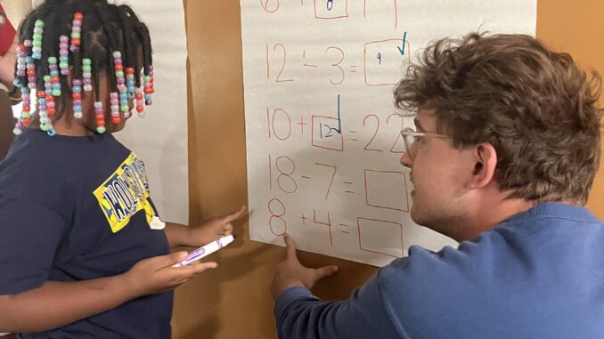 FEATURE A young man with short brown hair kneels and faces a poster paper. To the left of him is a young girl wearing box braids with beads, also facing the poster paper. On the paper itself is a series of addition and subtraction problems.