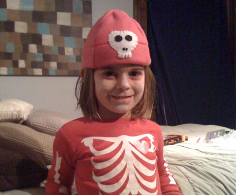 A young child with shoulder length brown hair smiles at the camera, wearing a pink long-sleeved shirt with images of bones on it, pink gloves with the same and a pink hat with a skeleton face sewn onto it. Behind them a bed with a gray comforter and several pillows is visible.