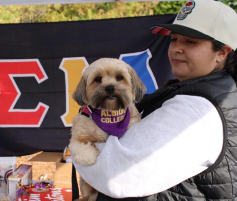 A woman in a baseball hat, gray sweatshirt and black puffy vest stands perpendicular to the camera holding a small light brown dog, who’s wearing a purple bandana around its neck which reads “Albion College.” Behind the pair is a black flag with colorful greek letters, and a table with food.