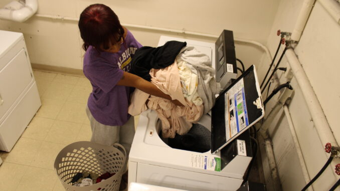Laundry A girl clad in oversized loungewear stands at a row of washing machines, arms full of various clothes being held over an open washing machine. The laundry room itself is bland but dusty, with a row of washers and dryers on opposite sides of the room, both white in color.