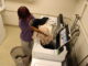 A girl clad in oversized loungewear stands at a row of washing machines, arms full of various clothes being held over an open washing machine. The laundry room itself is bland but dusty, with a row of washers and dryers on opposite sides of the room, both white in color.