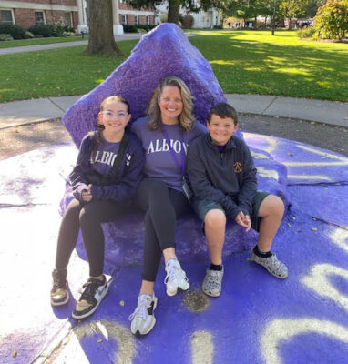 Three people sit against a purple spray-painted rock. In the background is a sidewalk and grassy lawn. On the left is a young girl wearing a purple t-shirt, leggings and golden Nike Dunks. In the middle is a middle-aged woman wearing a purple sweater, leggings and tennis shoes. On the right is a young boy wearing a dark purple pullover, shorts and Crocs. 