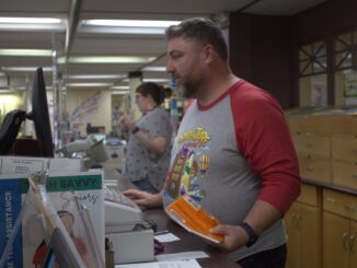 A man stands behind a long desk, looking at a computer screen and holding a book. Behind him are several cupboards and drawers, and in the background is another person standing behind the desk, speaking.