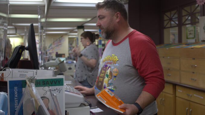 A man stands behind a long desk, looking at a computer screen and holding a book. Behind him are several cupboards and drawers, and in the background is another person standing behind the desk, speaking.