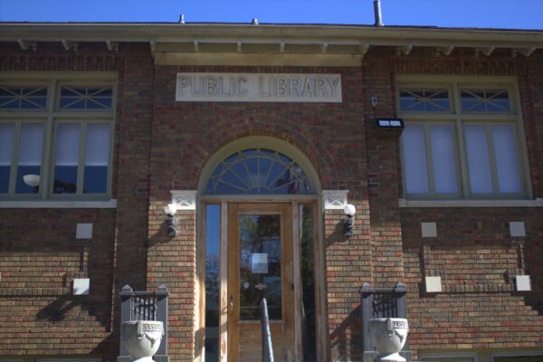 The front of a brick building with an ornate glass doorway and a fenced porch with ceramic planters on it. The brick is brown, and the sign above the single door reads “Public Library.”