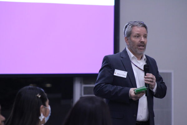 An older man stands to the right and in front of a purple projector screen. In the foreground are a few audience members looking toward the man, who is holding a small green box of cards and a mic.