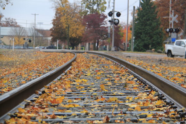 Curved railway tracks covered with golden autumn leaves continue into the horizon, surrounded by trees with vibrant fall foliage. A white truck is parked on the right.