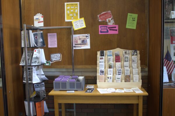 A table holds a display of small booklets of all different colors, and the wall behind it has a group of informational posters behind it.