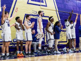 A line of seven basketball players stand and celebrate, many with their hands in the air. One coach sits on the bench and two stand beside the players. In the background is a purple wall with part of a golden “A” decal.