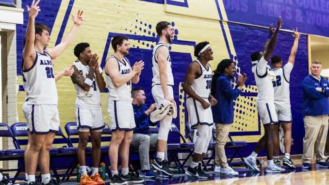 A line of seven basketball players stand and celebrate, many with their hands in the air. One coach sits on the bench and two stand beside the players. In the background is a purple wall with part of a golden “A” decal.