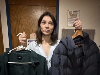 A young woman stands in front of the camera with a confused expression on her face. In each hand, she has a dark-colored coat on a hanger.