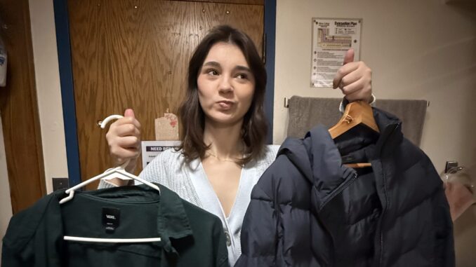A young woman stands in front of the camera with a confused expression on her face. In each hand, she has a dark-colored coat on a hanger.