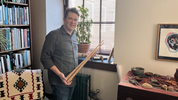 A man stands in front of a window holding two long wooden sticks used for harvesting rice.