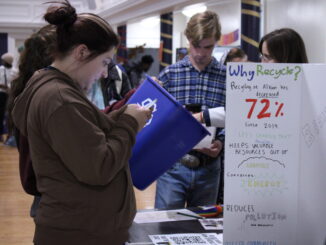 A young person stands in front of a table with papers on it while holding their phone out in front of them. In front of them is a tri-fold with one side visible, with words reading “Why recycle?” written in marker. In the background, there are more people standing and looking at other tables.