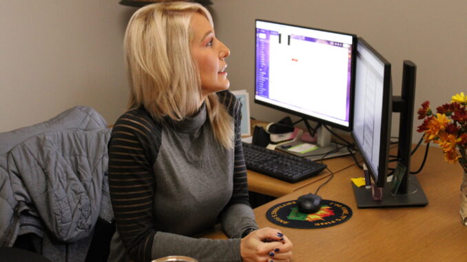 An adult woman sits up straight behind a wooden desk, her hands folded in front of her as she looks to the right, speaking. On the desk are two computer monitors and a vase of yellow and red flowers.