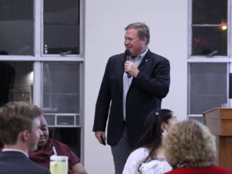 An adult man in semiformal wear speaks in front of an audience, holding a mic and smiling. Behind him are large windows looking out at a dark landscape.
