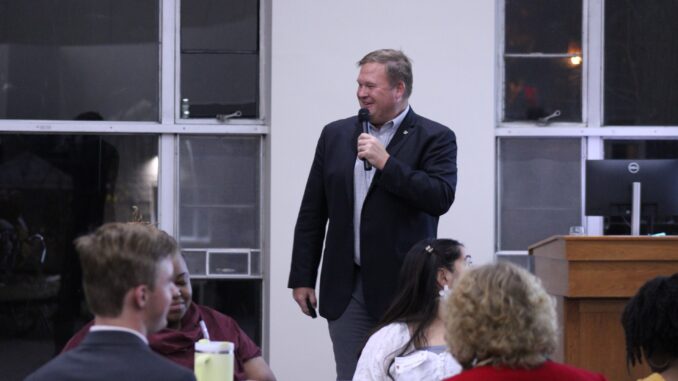 An adult man in semiformal wear speaks in front of an audience, holding a mic and smiling. Behind him are large windows looking out at a dark landscape.