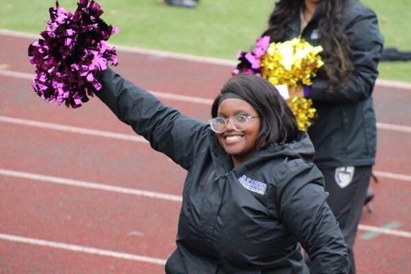 A young woman wearing glasses and a black jacket with the words “Albion cheerleading” written on it smiles at the camera while holding out a metallic purple pom pom. Behind her an outdoor track is visible, as well as another cheerleader holding metallic purple and gold pom poms.