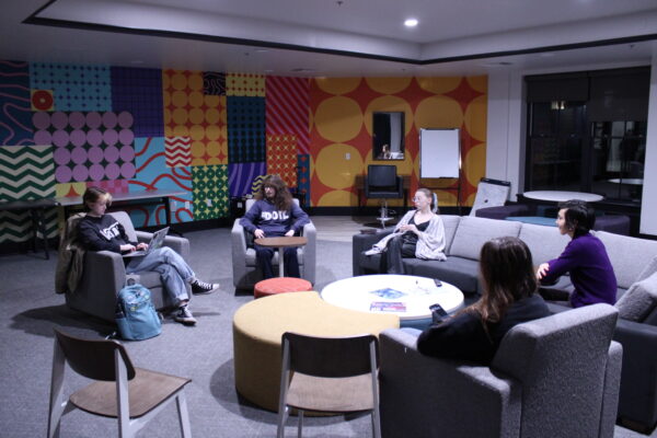 Five people gather in a room, all seated on light gray patterned chairs. Behind them is a multicolored and patterned wall, as well as a floor to ceiling window showing the outside. In front of one of the individuals is a small oval table with a singular white leg.