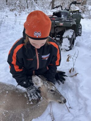 A girl sits in the snow with a deer laying across her lap as she holds its head up. The ground beneath her has a layer of snow on it, and there is a four-wheeler in the background.