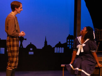 A young adult wearing plaid pants and suspenders stands to the left of the frame, perpendicular to the camera, smiling at a young woman sitting on a wooden bench to the right of the frame. She sits in a navy and white dress, body angled towards the camera and head turned to her right, looking up and smiling at the other person. They’re on a wood-paneled stage, in the background is a dark blue screen with the black silhouettes of buildings.