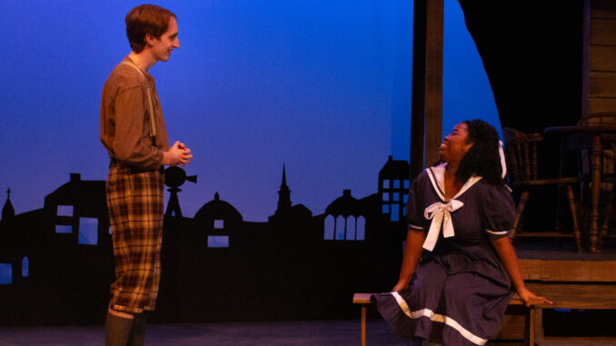 A young adult wearing plaid pants and suspenders stands to the left of the frame, perpendicular to the camera, smiling at a young woman sitting on a wooden bench to the right of the frame. She sits in a navy and white dress, body angled towards the camera and head turned to her right, looking up and smiling at the other person. They’re on a wood-paneled stage, in the background is a dark blue screen with the black silhouettes of buildings.