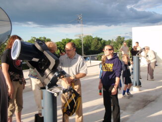 A group of people stand in front of an outdoor telescope or around a platform. The location is outside, and the floor beneath the people is white and has a tarplike texture.