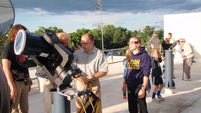 A group of people stand in front of an outdoor telescope or around a platform. The location is outside, and the floor beneath the people is white and has a tarplike texture.