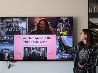 A TV screen showing scenes and posters from movies and TV series created by horror director Mike Flanagan. In the middle of the images, over a red background, are the words “A Complete Guide to the Mike Flana-verse.” A young adult stands next to the screen, looking at it and pointing at the poster for the series “The Midnight Club.”