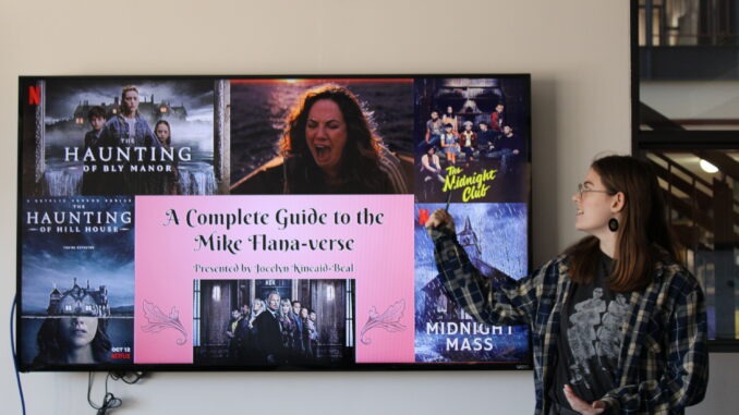 A TV screen showing scenes and posters from movies and TV series created by horror director Mike Flanagan. In the middle of the images, over a red background, are the words “A Complete Guide to the Mike Flana-verse.” A young adult stands next to the screen, looking at it and pointing at the poster for the series “The Midnight Club.”